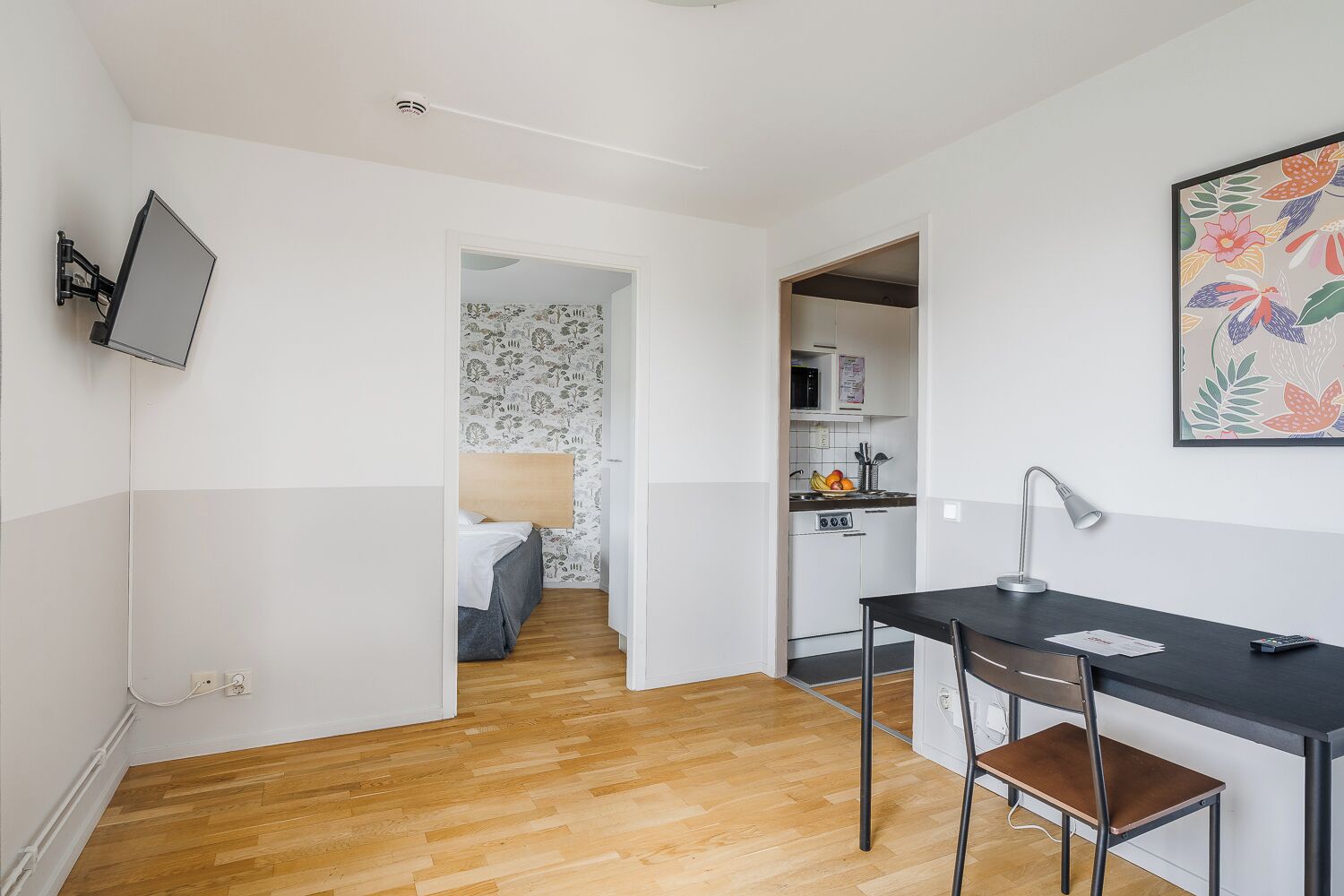 Una habitación compacta con cama, escritorio y silla, televisor montado en la pared y zona de cocina.