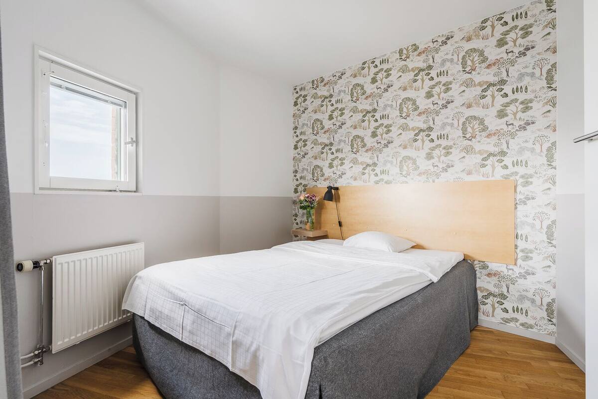 A bedroom with a bed, a radiator, a window, and a wall with a nature-themed wallpaper.