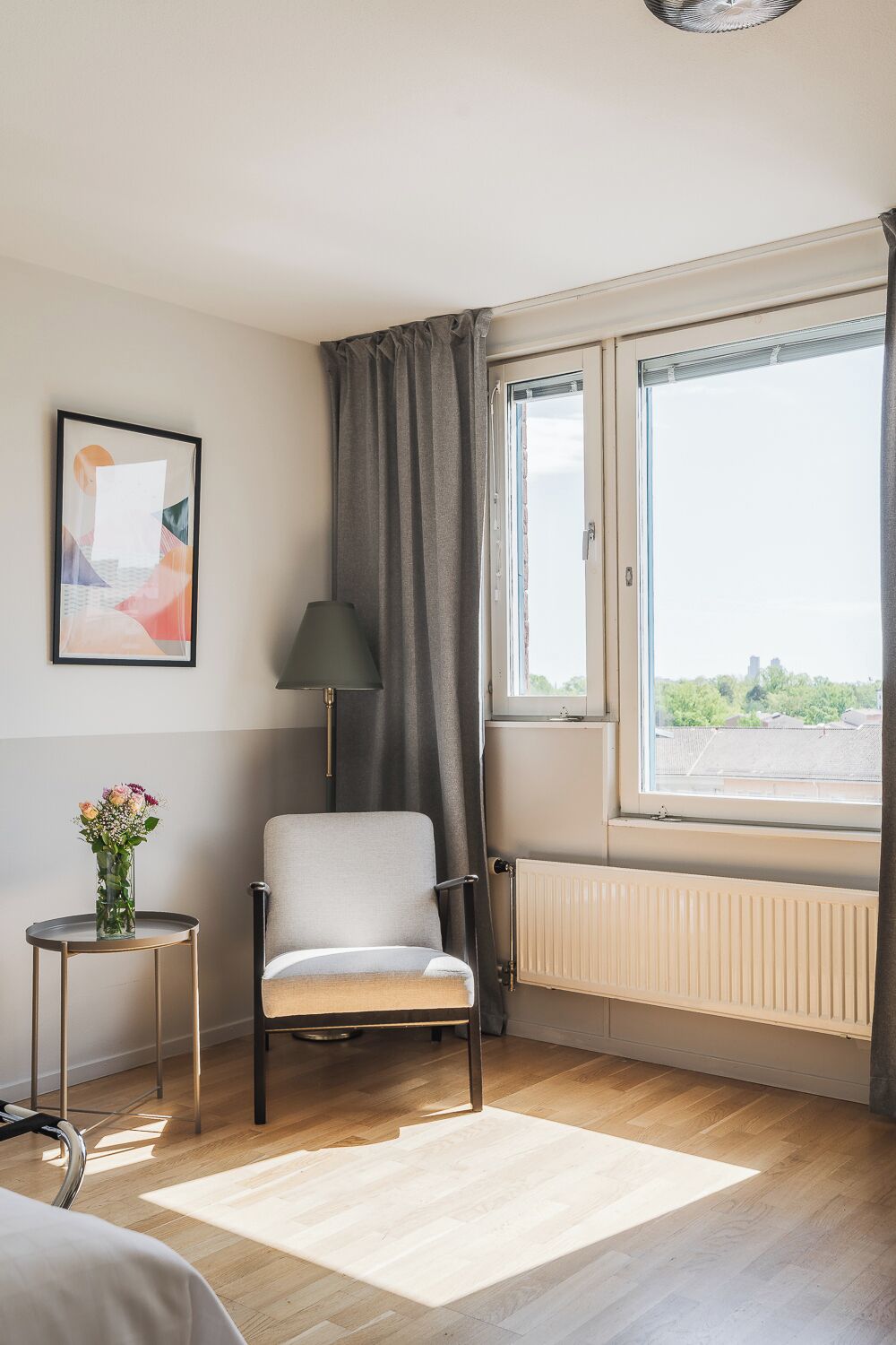 A room with a chair, a radiator, a window with a view, a lamp, and a framed picture on the wall.