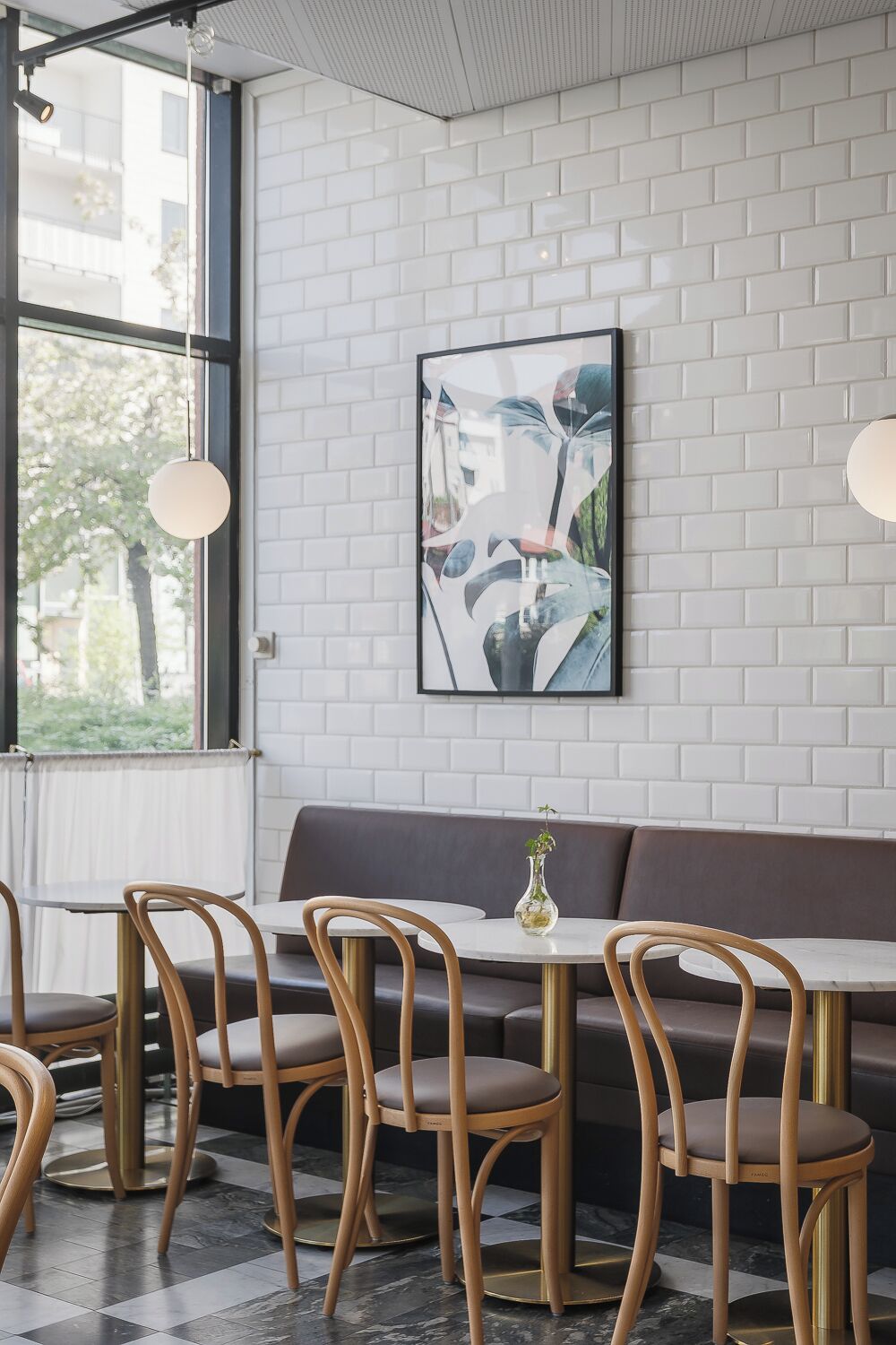 Interior de restaurante con pared de ladrillo blanco, un ventanal grande, un cuadro abstracto enmarcado, un banco acolchado marrón y mesas redondas con sillas de madera.