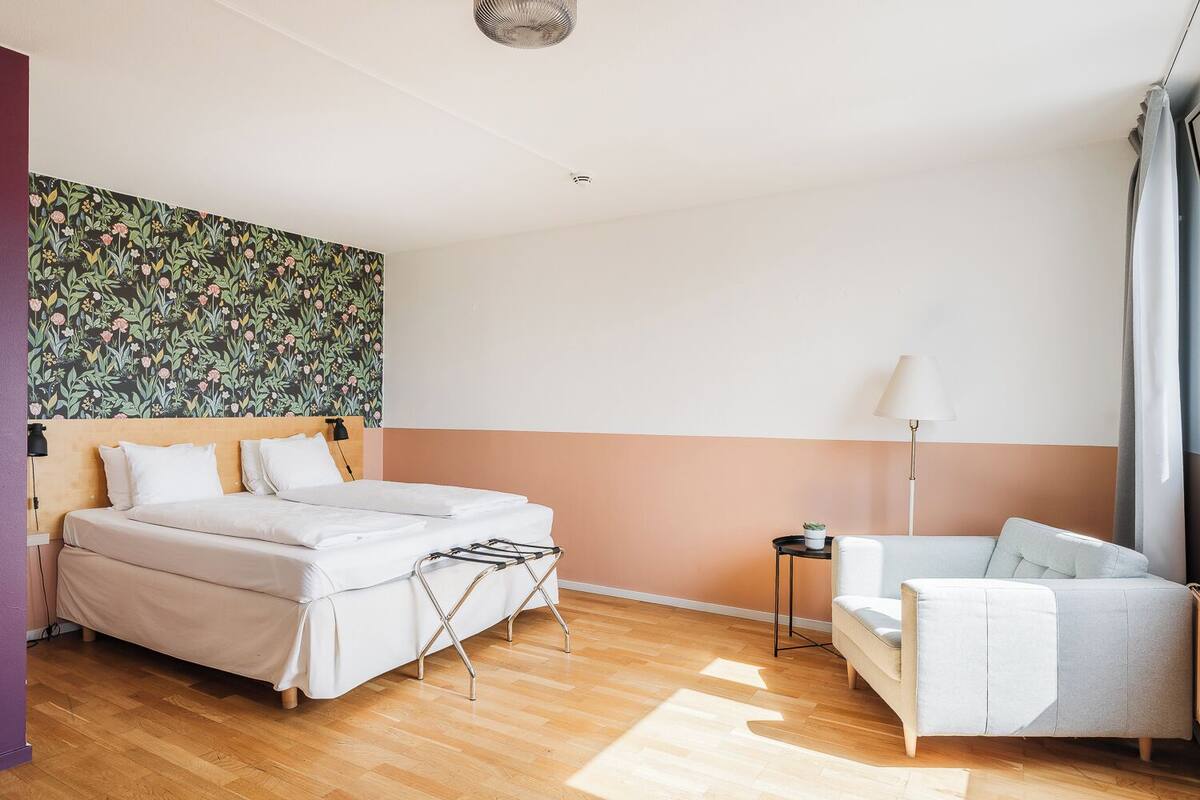 A hotel room with a bed, a small table, a chair, a floor lamp, and a wall with a floral pattern.