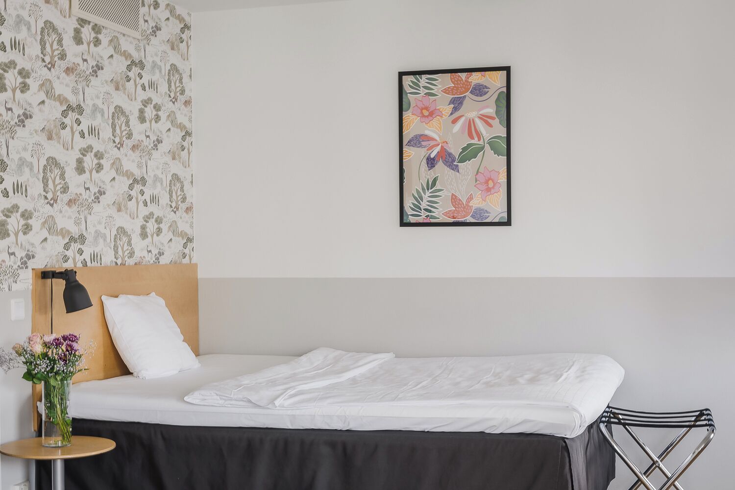 Uma cama bem arrumada com lençóis brancos, uma cadeira de metal preta, uma mesinha redonda com um vaso de flores e uma parede com uma pintura floral.
