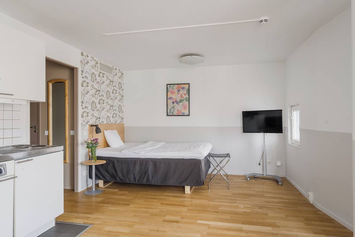 A compact hotel room with a bed, a small table with a vase, a TV, and a kitchenette.