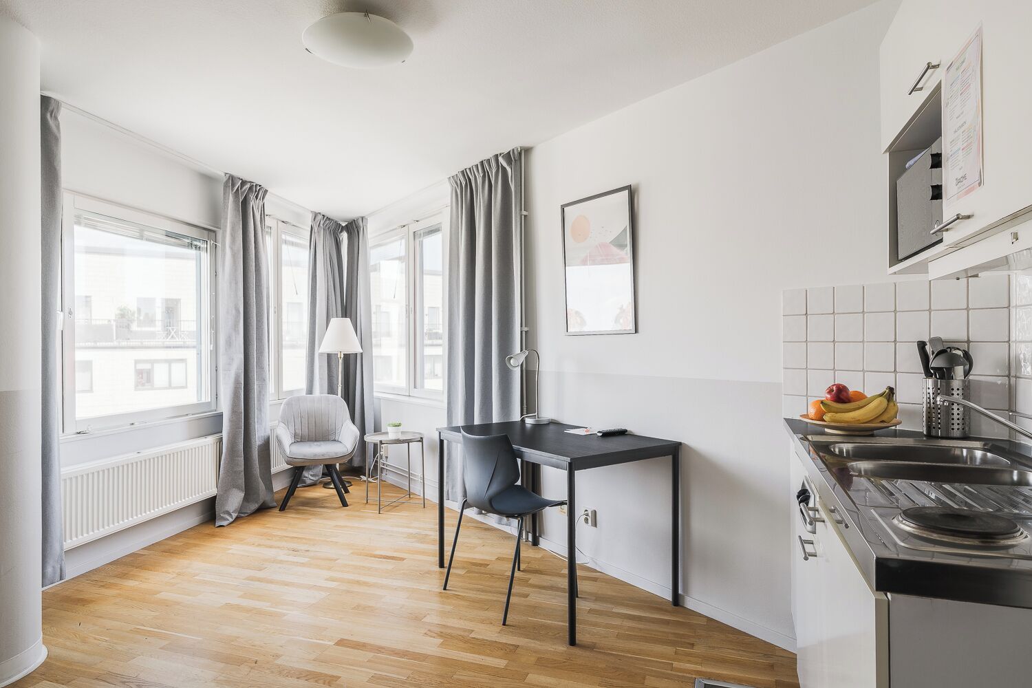 A compact kitchen with a dining area, a radiator, a window with curtains, and a sink with a fruit bowl.