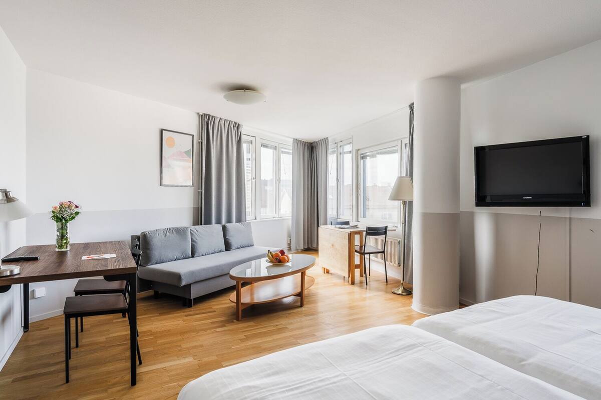 A hotel room with a bed, a sofa, a desk with a lamp, a coffee table, and a TV.