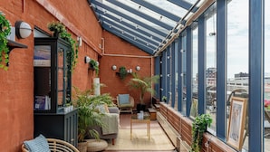 A covered balcony with a glass ceiling, potted plants, a seating area with a sofa and chairs, and a view of the cityscape. - 2Home Hotel Apartments (Solna)