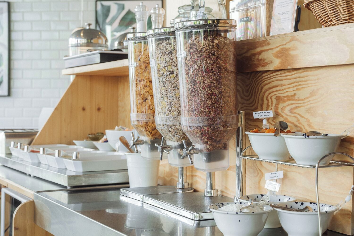 A food station with dispensers for granola and dried fruits, a coffee machine, and a shelf with bowls of yogurt and other dairy products.