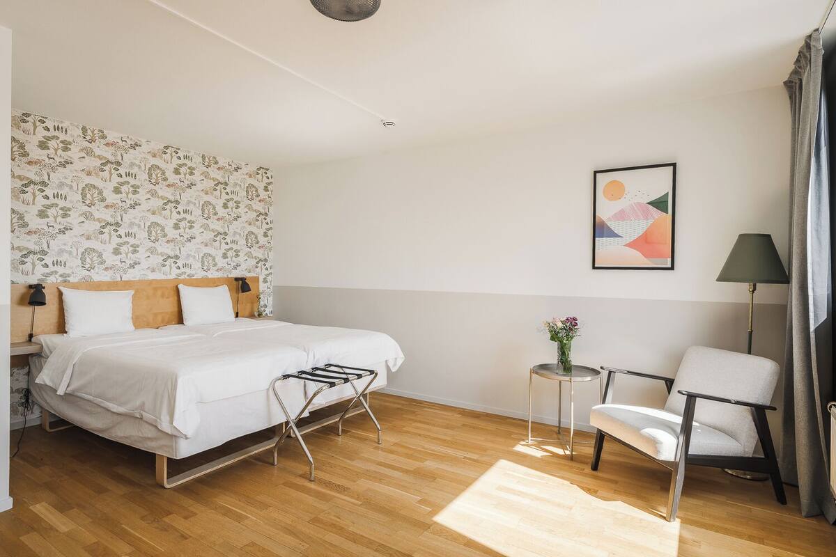 A bedroom with a bed, a chair, a small table with flowers, and a wall with a patterned wallpaper.