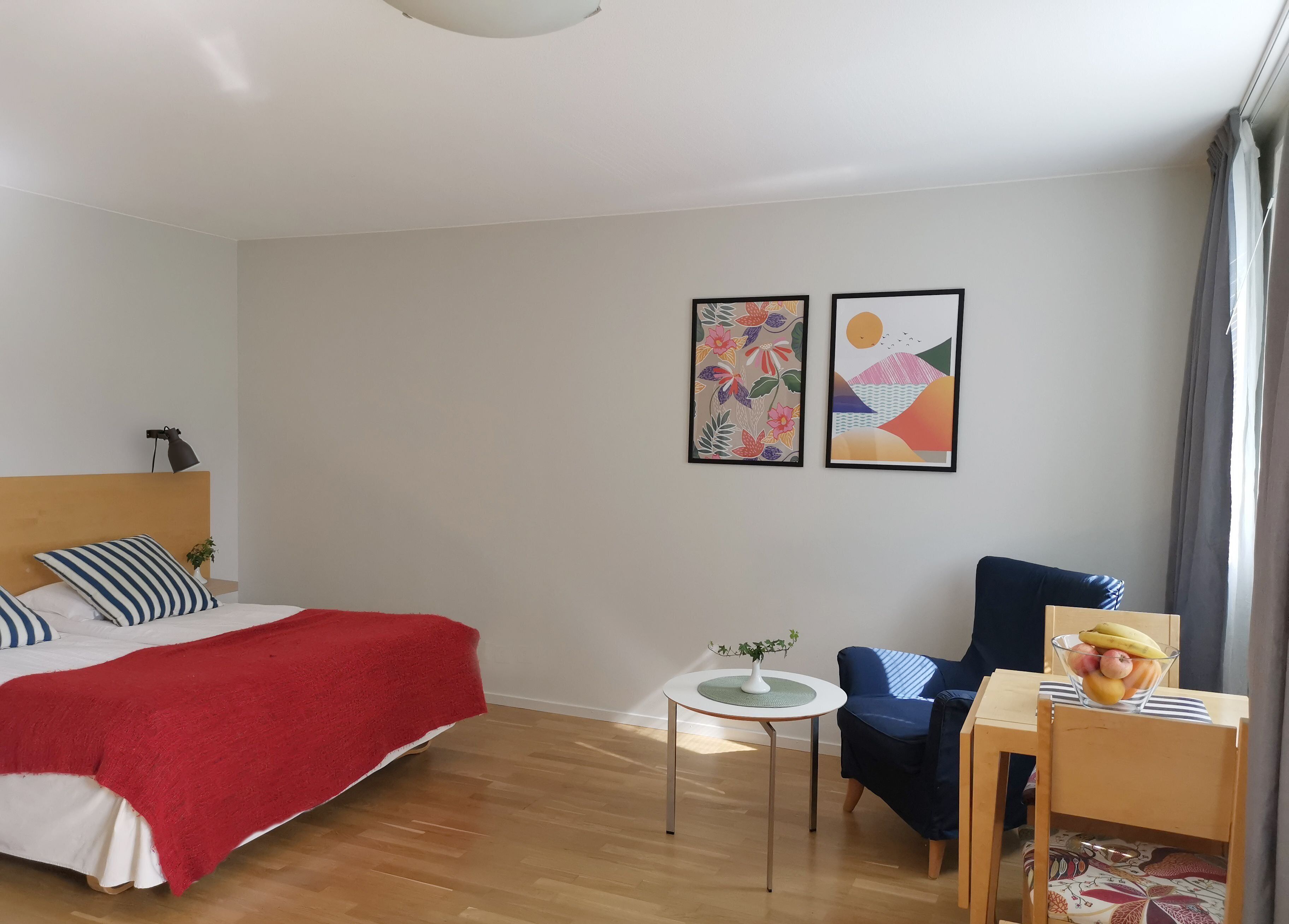 Une chambre d’hôtel moderne avec un lit en bois, une couverture rouge, une petite table ronde ornée d’un vase, une chaise bleue et deux œuvres d’art encadrées accrochées au mur.