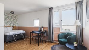 A room with a bed, a desk, a chair, a small table with a plant, and a view of the city. - 2Home Hotel Apartments (Solna)