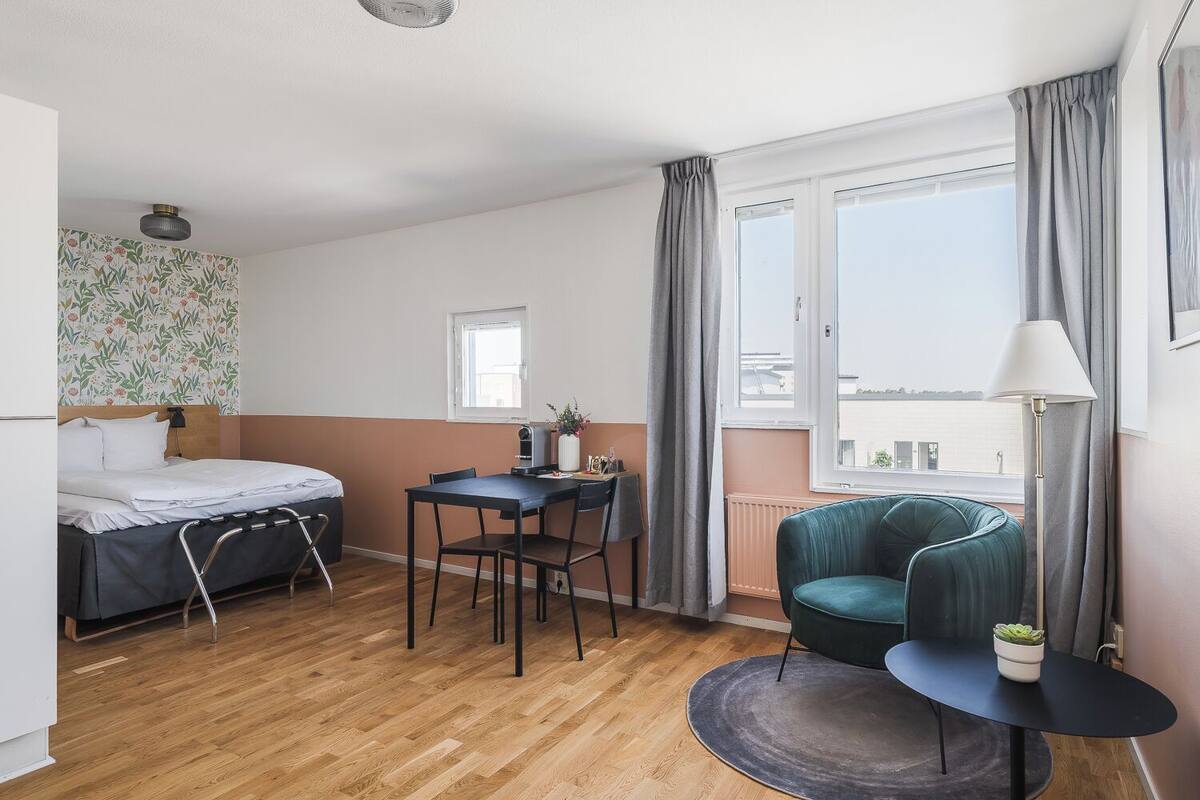 A room with a bed, a desk, a chair, a small table with a plant, and a view of the city.
