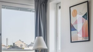 A room with a teal chair, a small table with a plant, a lamp, and a city view through a window. - 2Home Hotel Apartments (Solna)