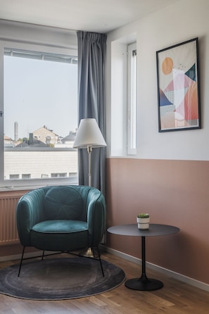 A room with a teal chair, a small table with a plant, a lamp, and a city view through a window. - 2Home Hotel Apartments (Solna)