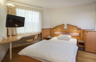 A hotel room with a bed, a desk with a television, a chair, a nightstand, and a wooden headboard.