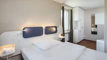 A modern hotel room with a large bed, a bedside table with a phone, a mirror, and a small desk with a lamp.