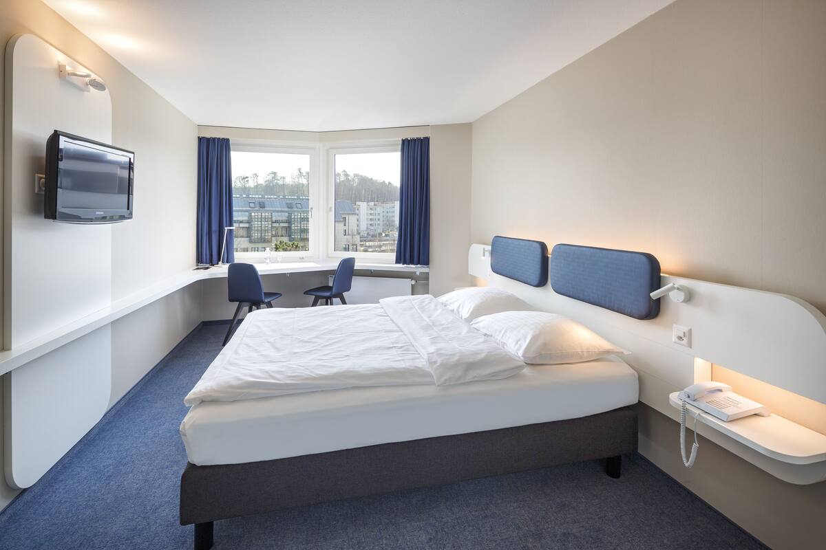 A hotel room with a bed, a desk, a chair, a TV, and a window with curtains.