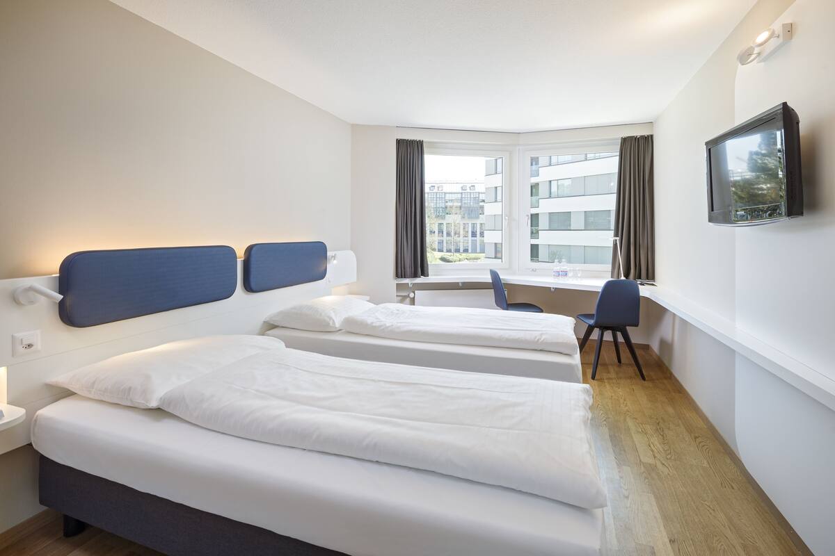 A hotel room with two beds, a desk, a chair, a TV, and a window with curtains.