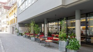 Een terras met tafels en stoelen, plantenpotten en een gebouw met grote ramen op de achtergrond.