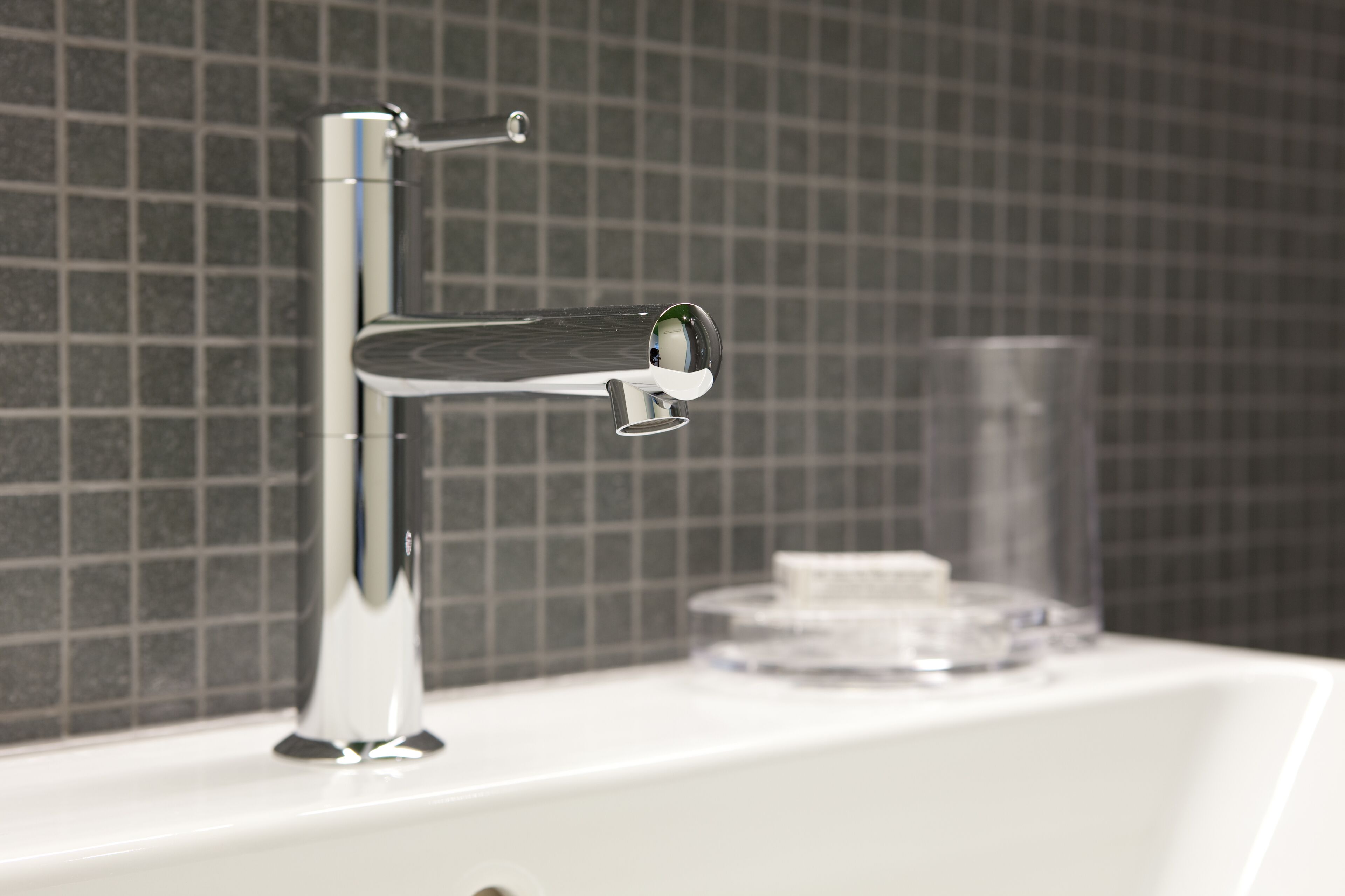 A modern bathroom with a chrome faucet, a clear glass container, and a tiled wall.