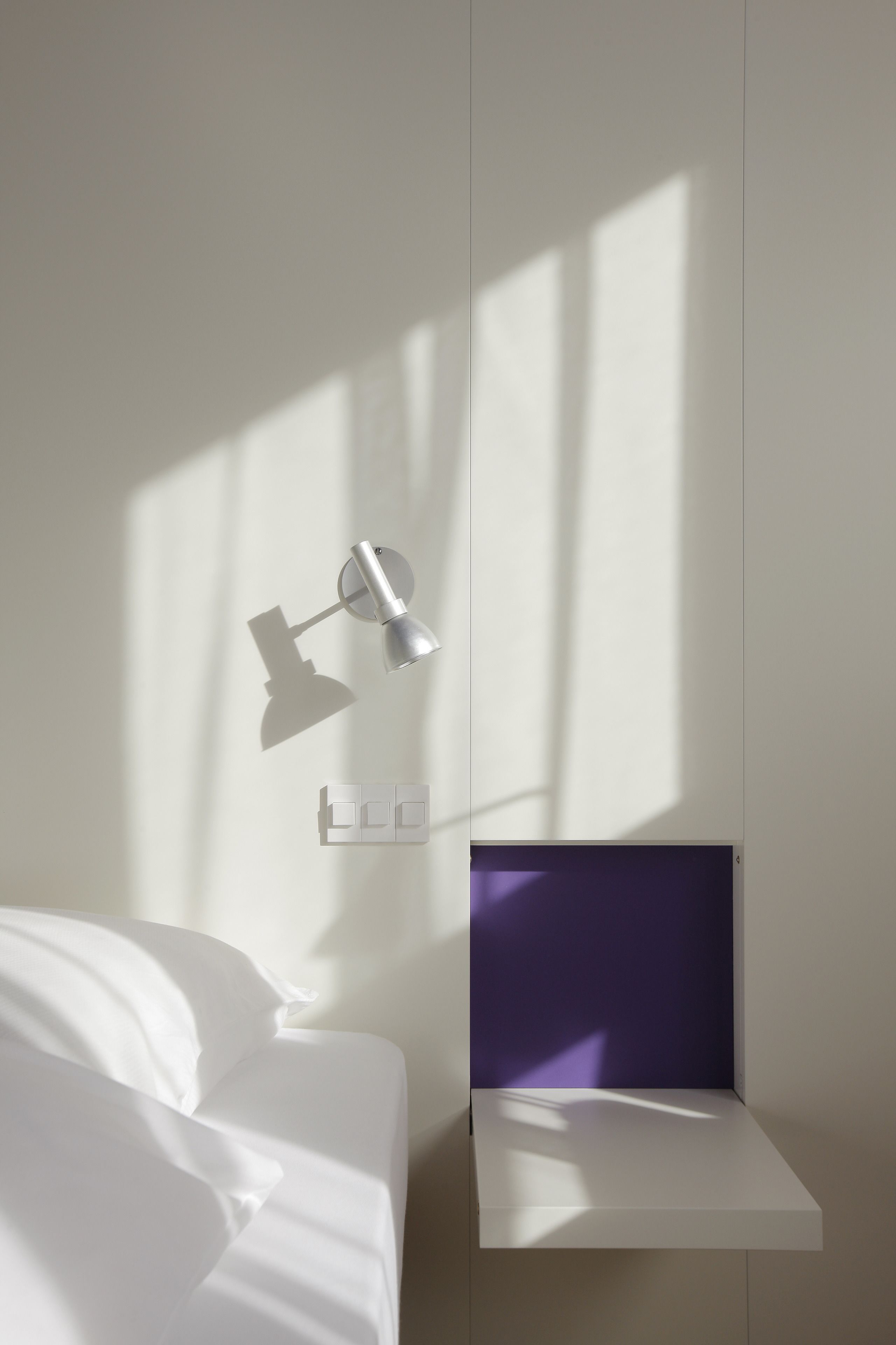 Ein schlichter Raum mit einem Bett, einer Wandlampe und einer kleinen Ablagefläche mit einem Bildschirm.