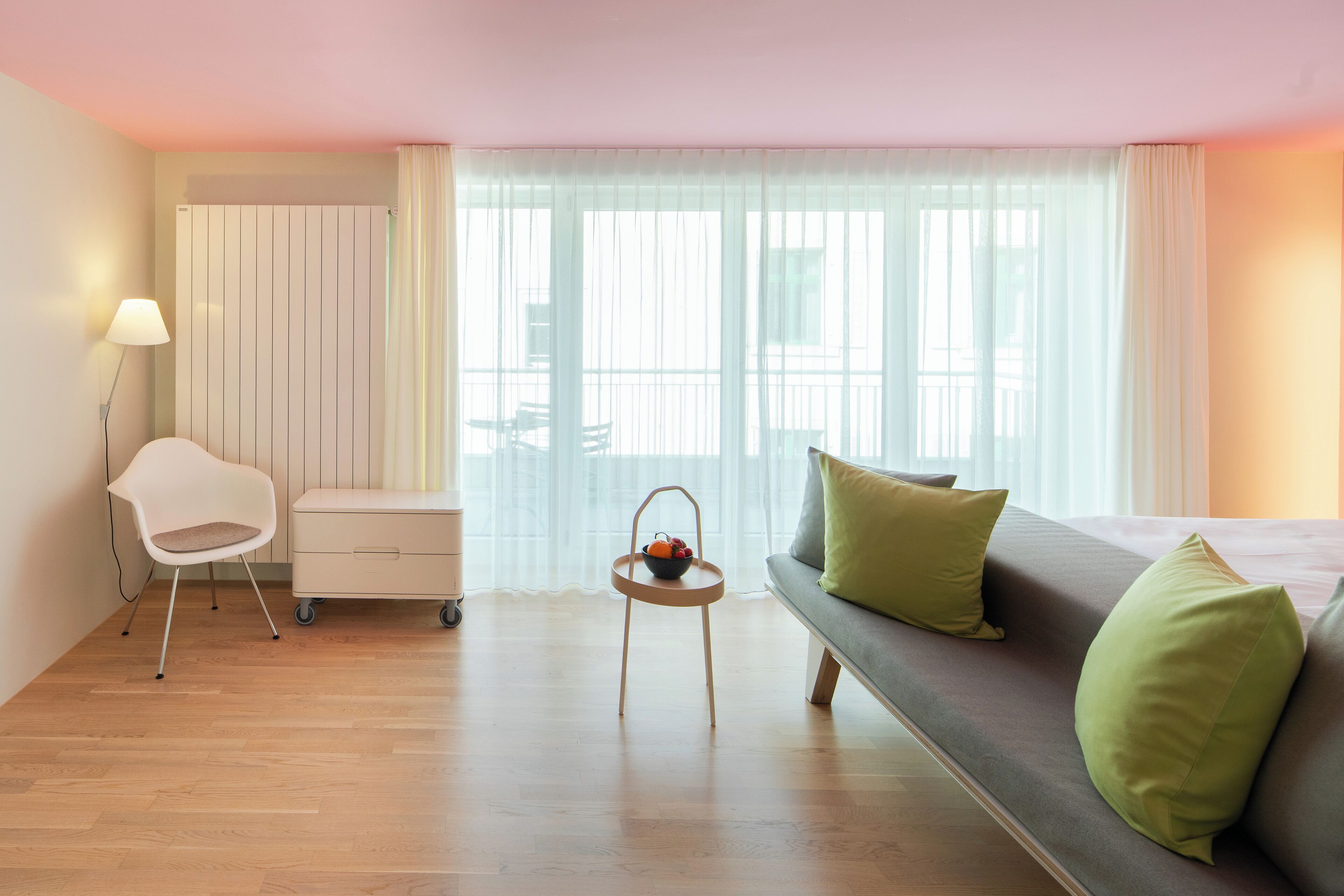 A modern hotel room with a wooden floor, a grey sofa with green cushions, a white chair, a small table with a fruit bowl, and a large window with sheer curtains.