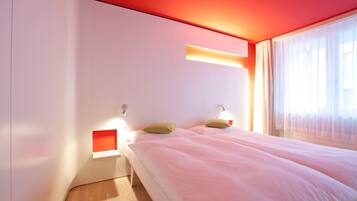 Una habitación de hotel moderna con una cama grande, dos mesitas de noche y un ventanal con cortinas de gasa.