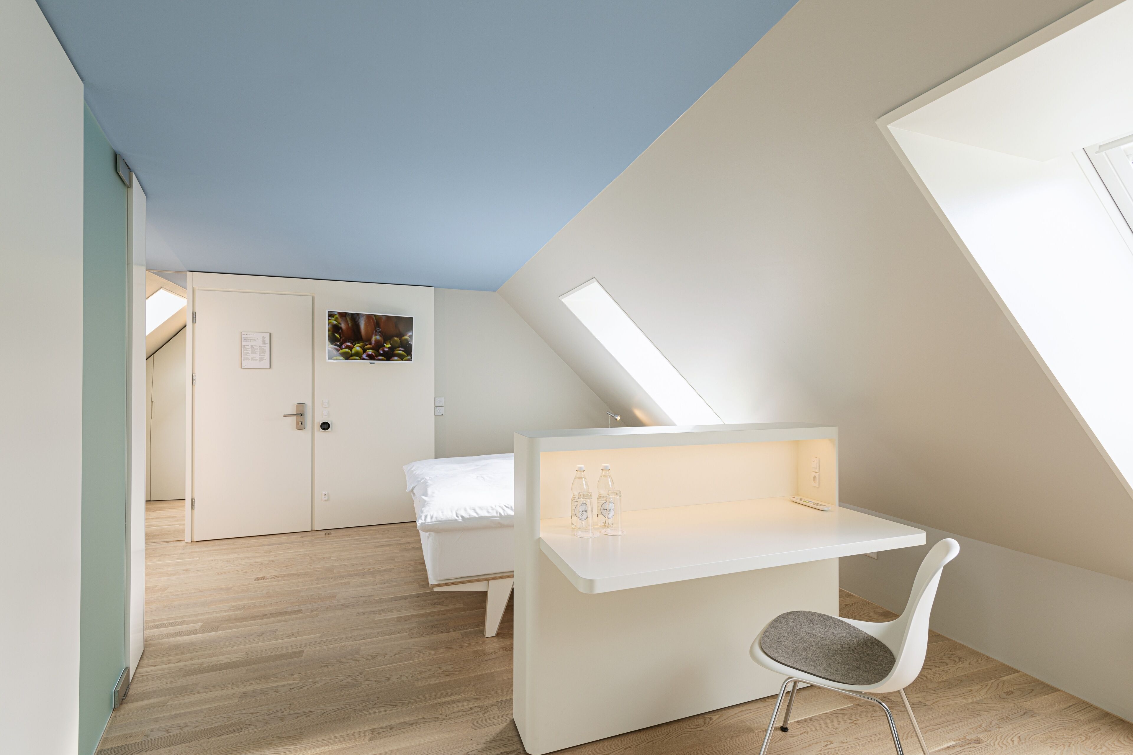 A modern, minimalist bedroom with a sloped ceiling, a built-in desk, and a single bed.