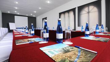 Un salón de conferencias con alfombra roja, varias mesas con botellas de agua y vasos, y una pantalla para proyector.