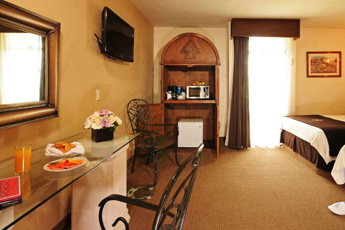 Habitación de hotel con cama, escritorio con desayuno, televisor y kitchenette pequeña.