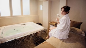 A room with a large bathtub, a bed with pillows, and a small table with a glass of water.