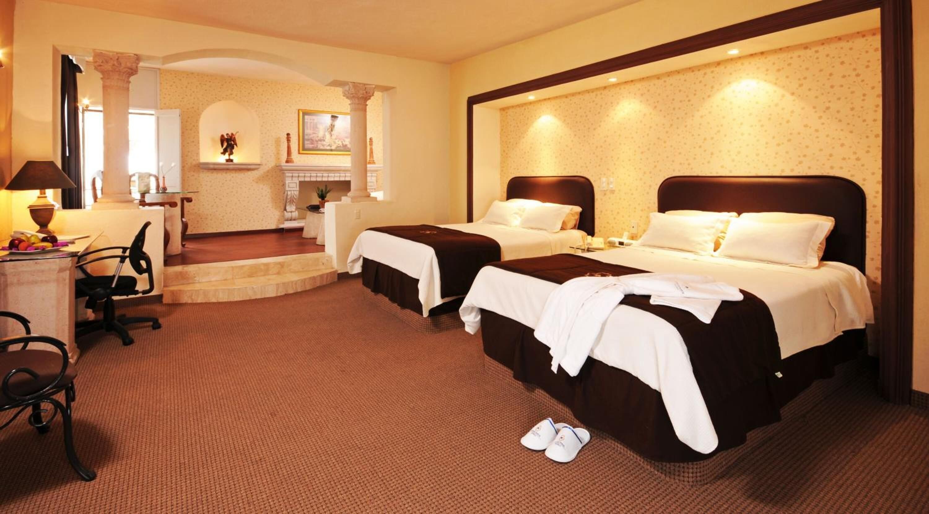 A hotel room with two beds, a desk, and a chair. There is a fireplace and a small statue on the mantel.
