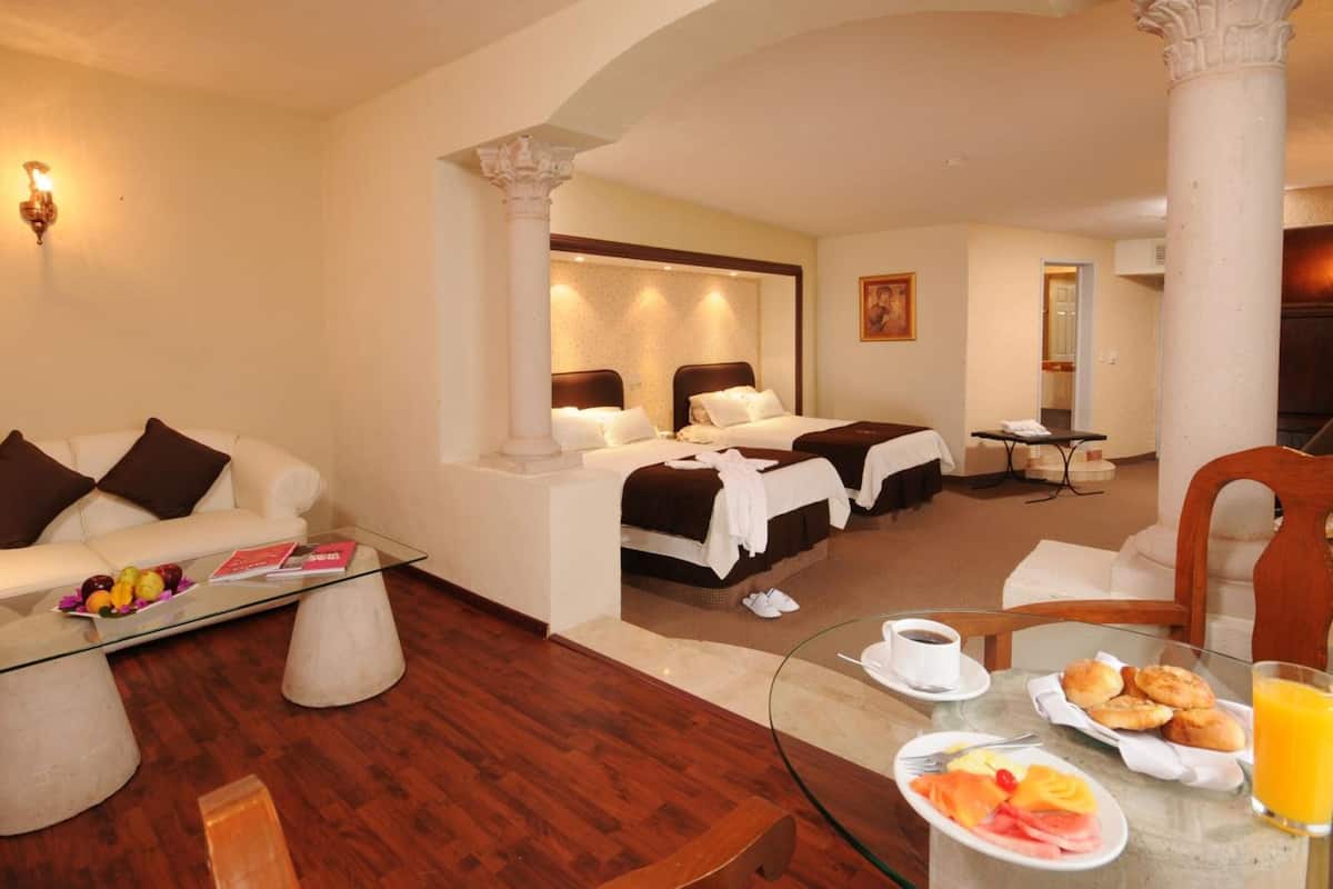 Habitación de hotel con sofá, dos camas, una mesa de centro de vidrio y alimentos para el desayuno sobre una mesa.