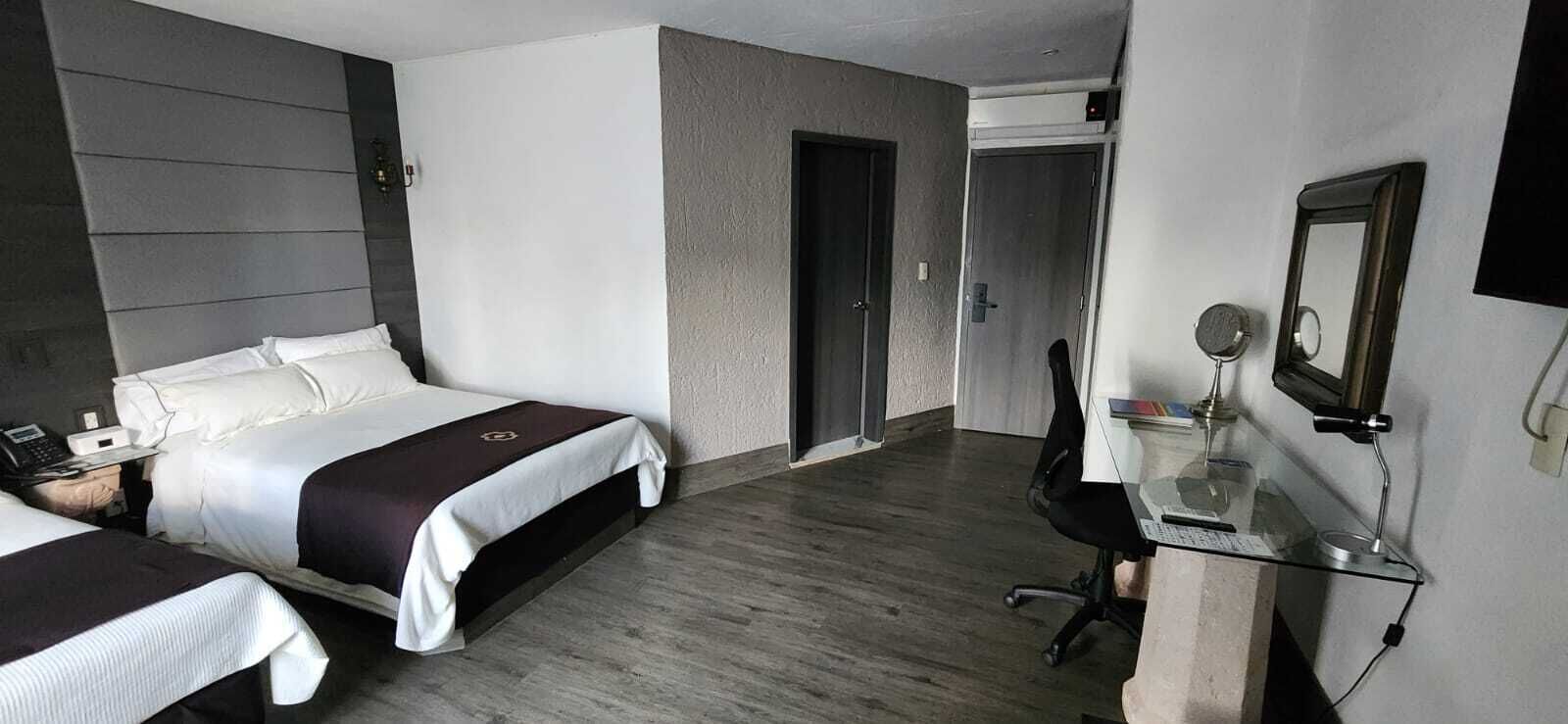 A hotel room with two beds, a desk with a chair, a mirror, and a door leading to another room.