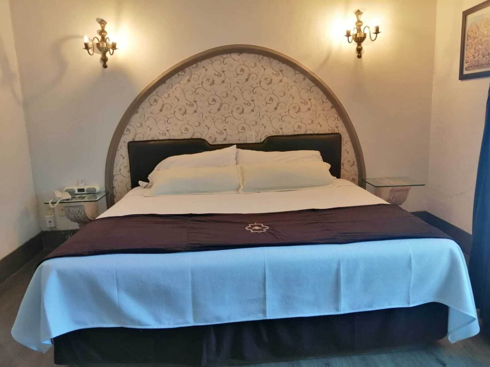 Ein ordentlich bezogenes Bett mit dunklem Kopfteil und einer dekorativen Wandverkleidung darüber. An jeder Seite des Bettes befinden sich zwei Wandleuchten.
