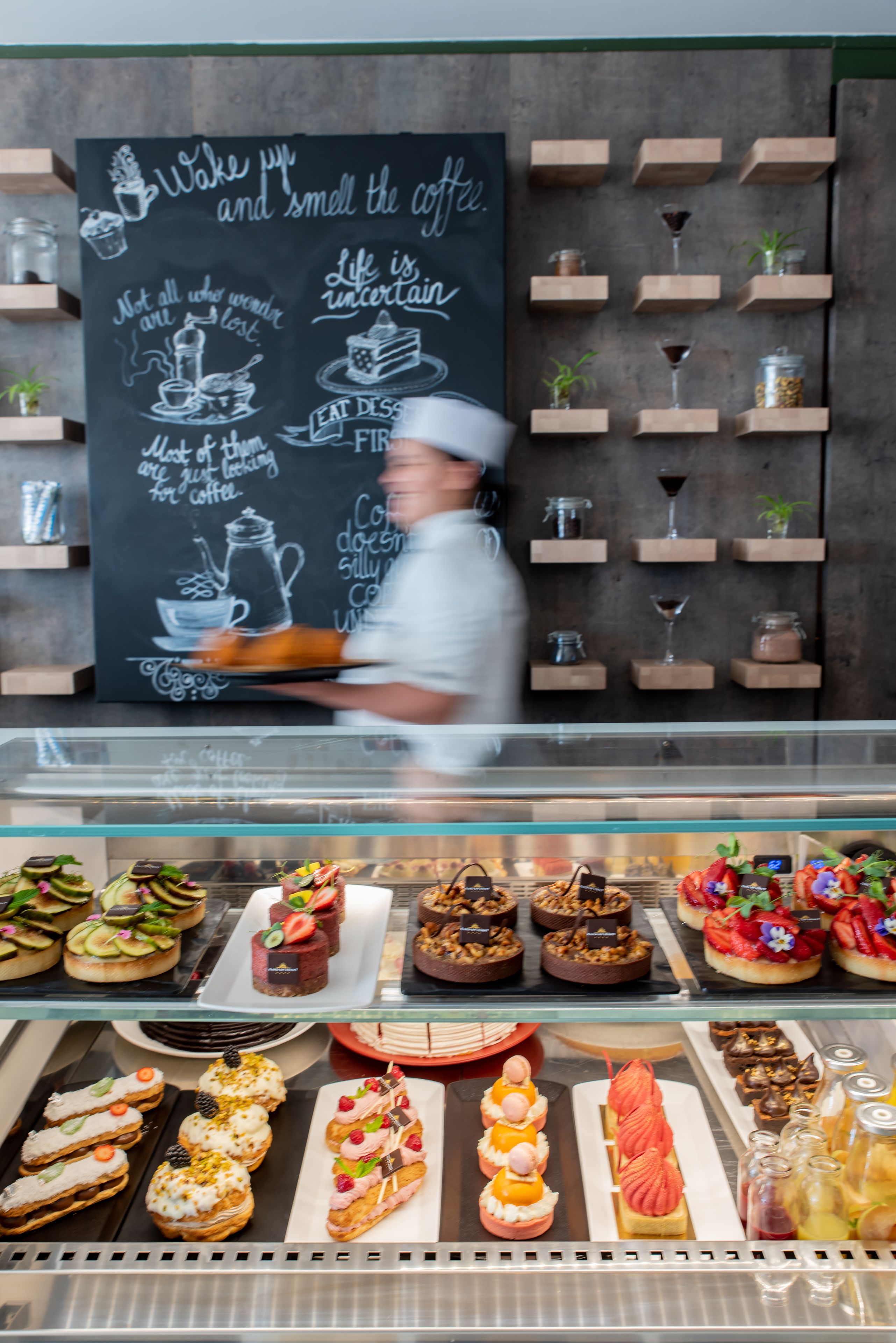 A bakery display case with various pastries and desserts, a chalkboard with motivational quotes, and shelves with jars and glasses.