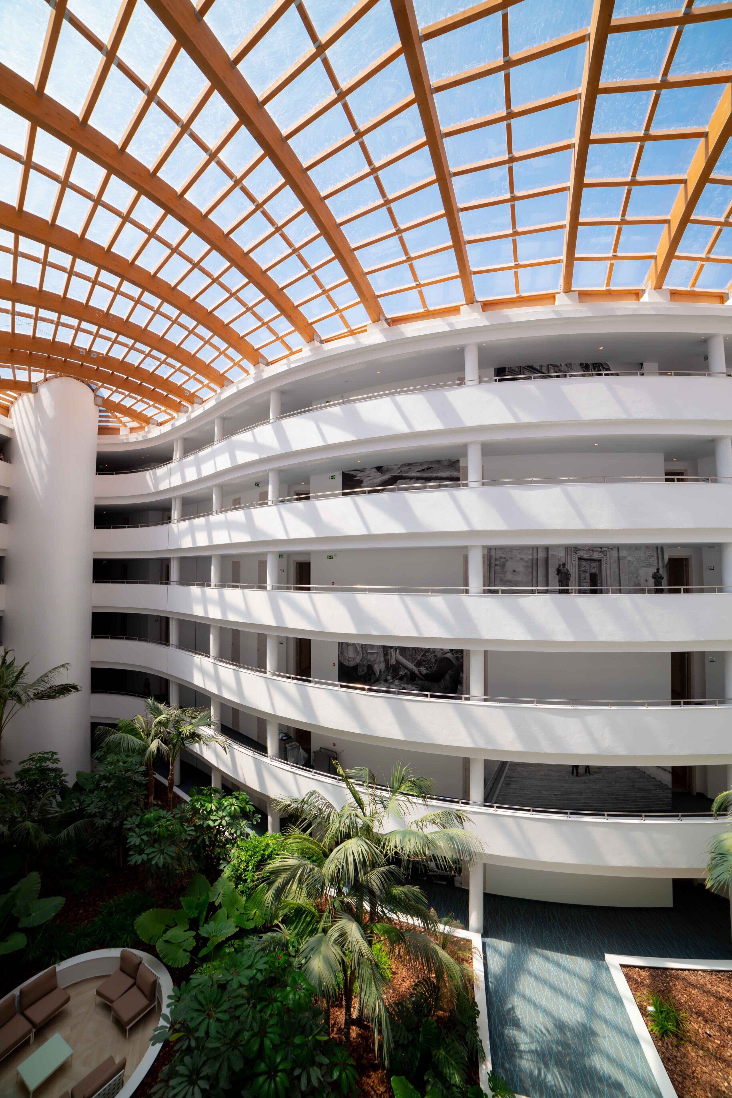 A multi-level atrium with a glass ceiling, indoor plants, and a central seating area.