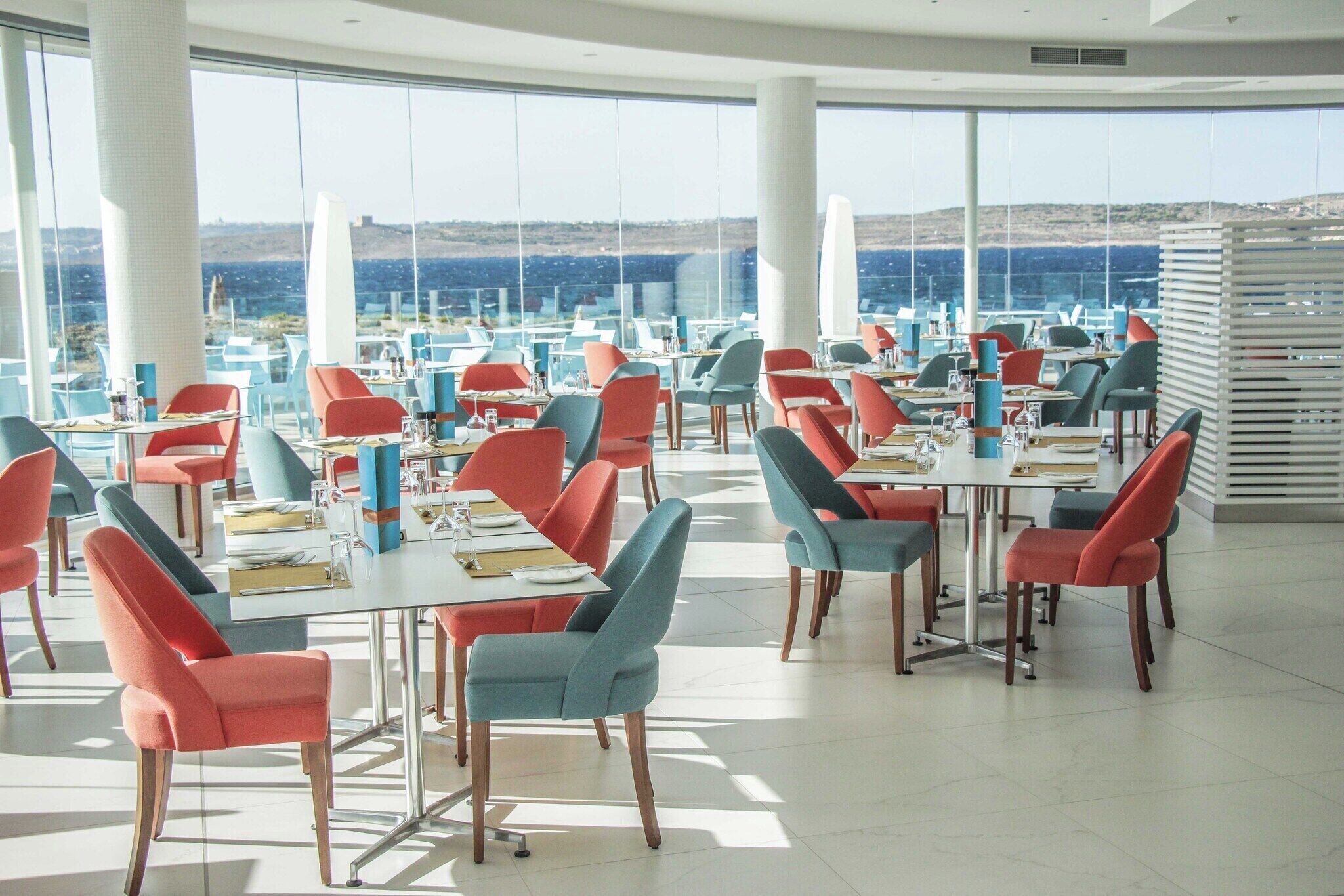 A spacious dining area with modern chairs and tables, large windows offering a view of the sea and hills.