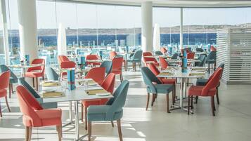 A spacious dining area with modern chairs and tables, large windows offering a view of the sea and hills.
