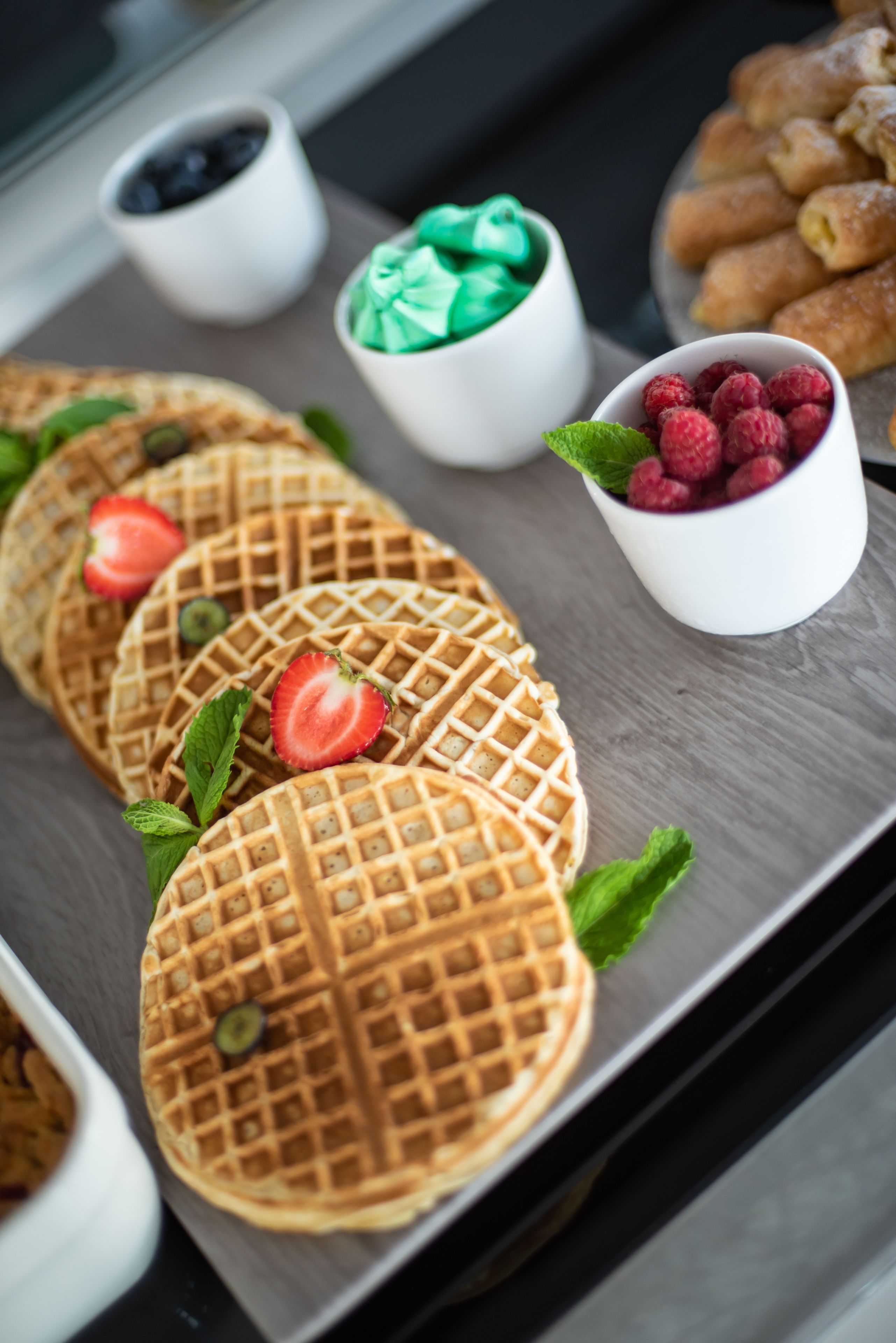 A tray of waffles with strawberries and mint leaves, accompanied by a cup of raspberries and a small bowl of green dessert.