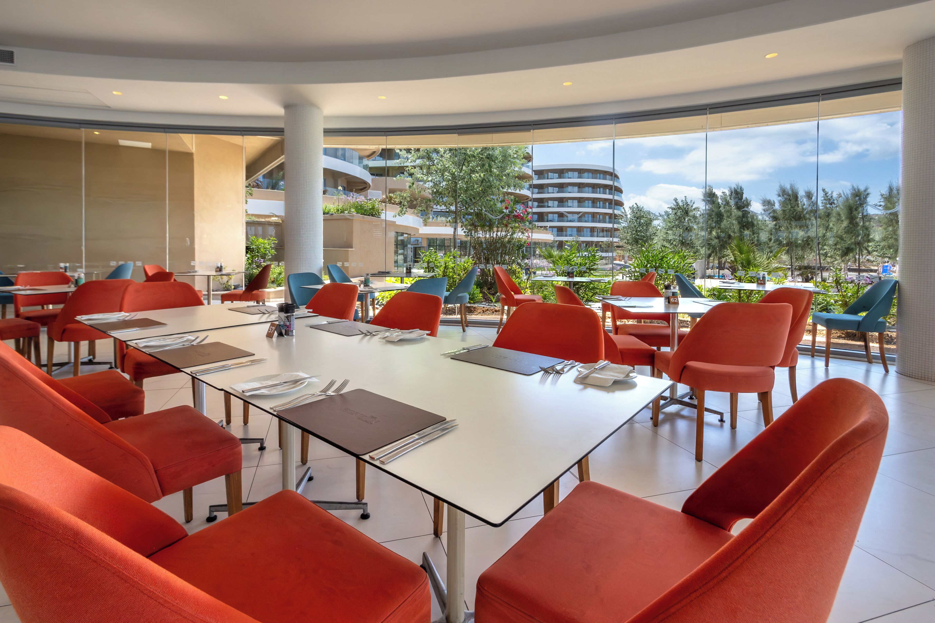 A modern dining area with red chairs, white tables, and large windows offering a view of buildings and trees.
