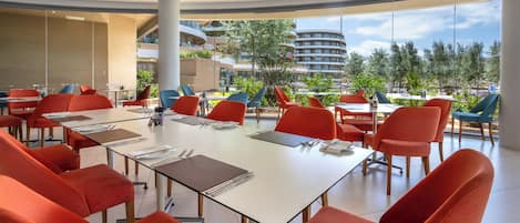 A modern dining area with red chairs, white tables, and large windows offering a view of buildings and trees.