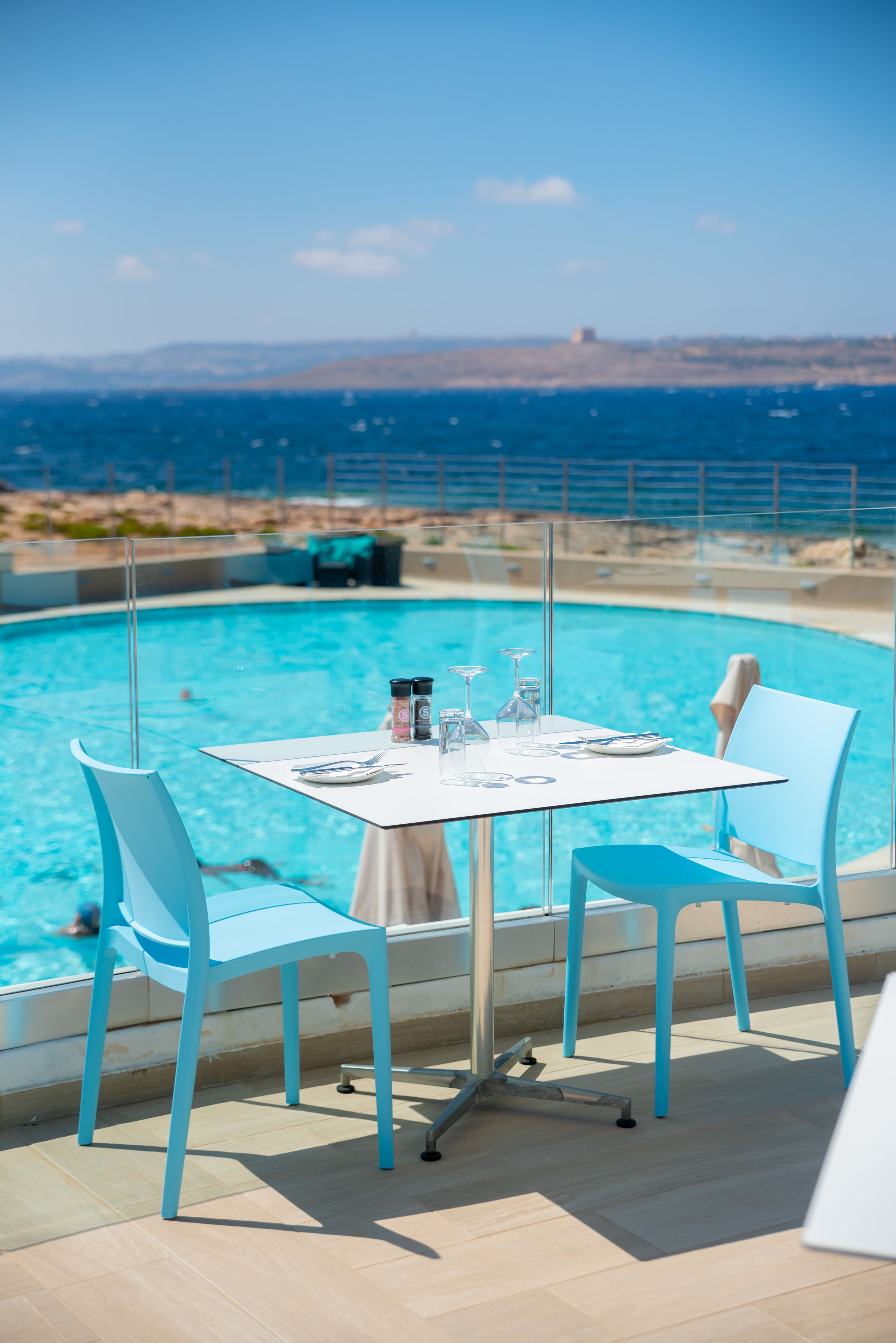 Un espace repas aménagé au bord de la piscine, avec une table dressée pour deux personnes, des chaises et une vue sur la mer.