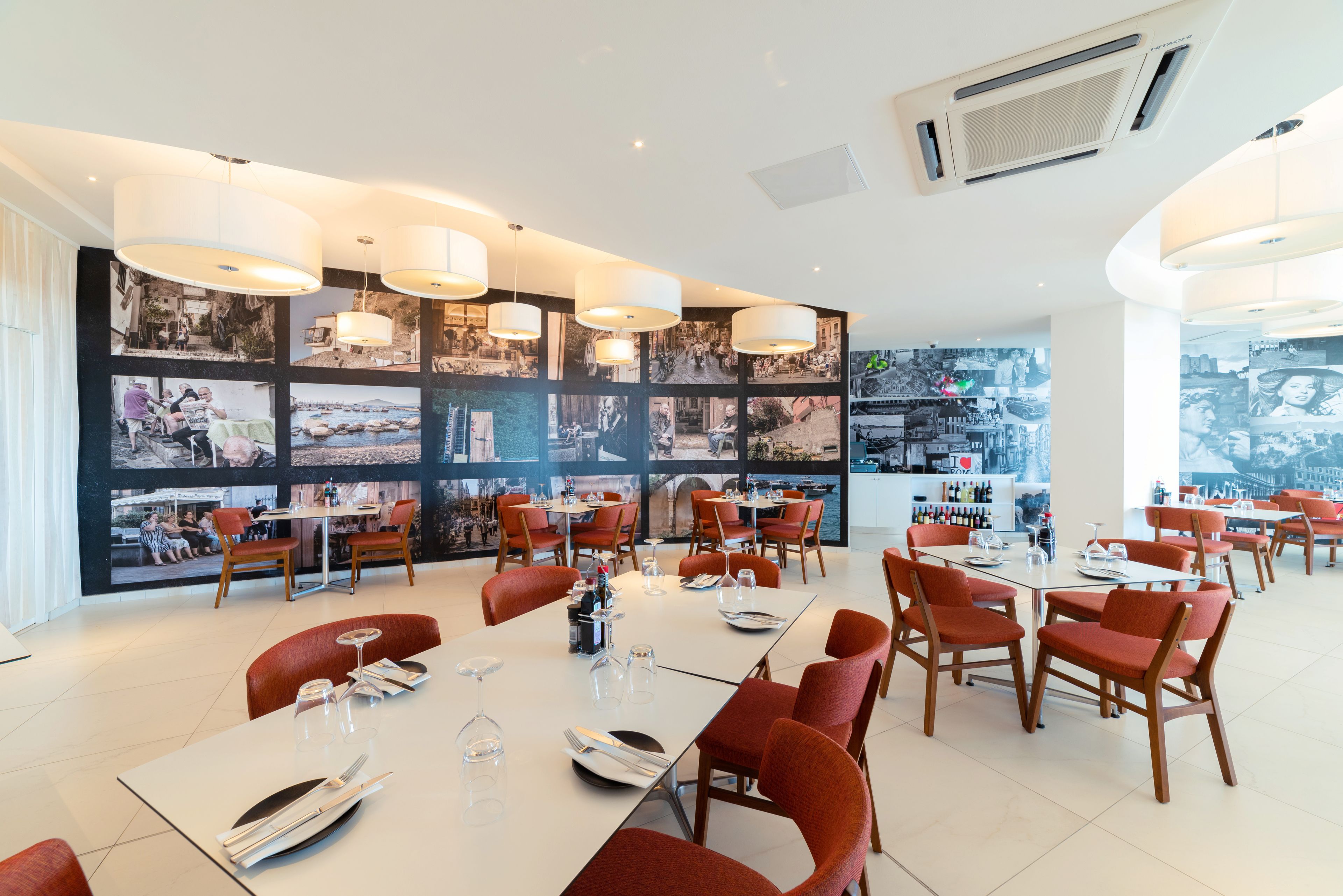 Un comedor moderno con varias mesas y sillas, una pared con diversas fotografías, y vistas de la ciudad a través de amplias ventanas.