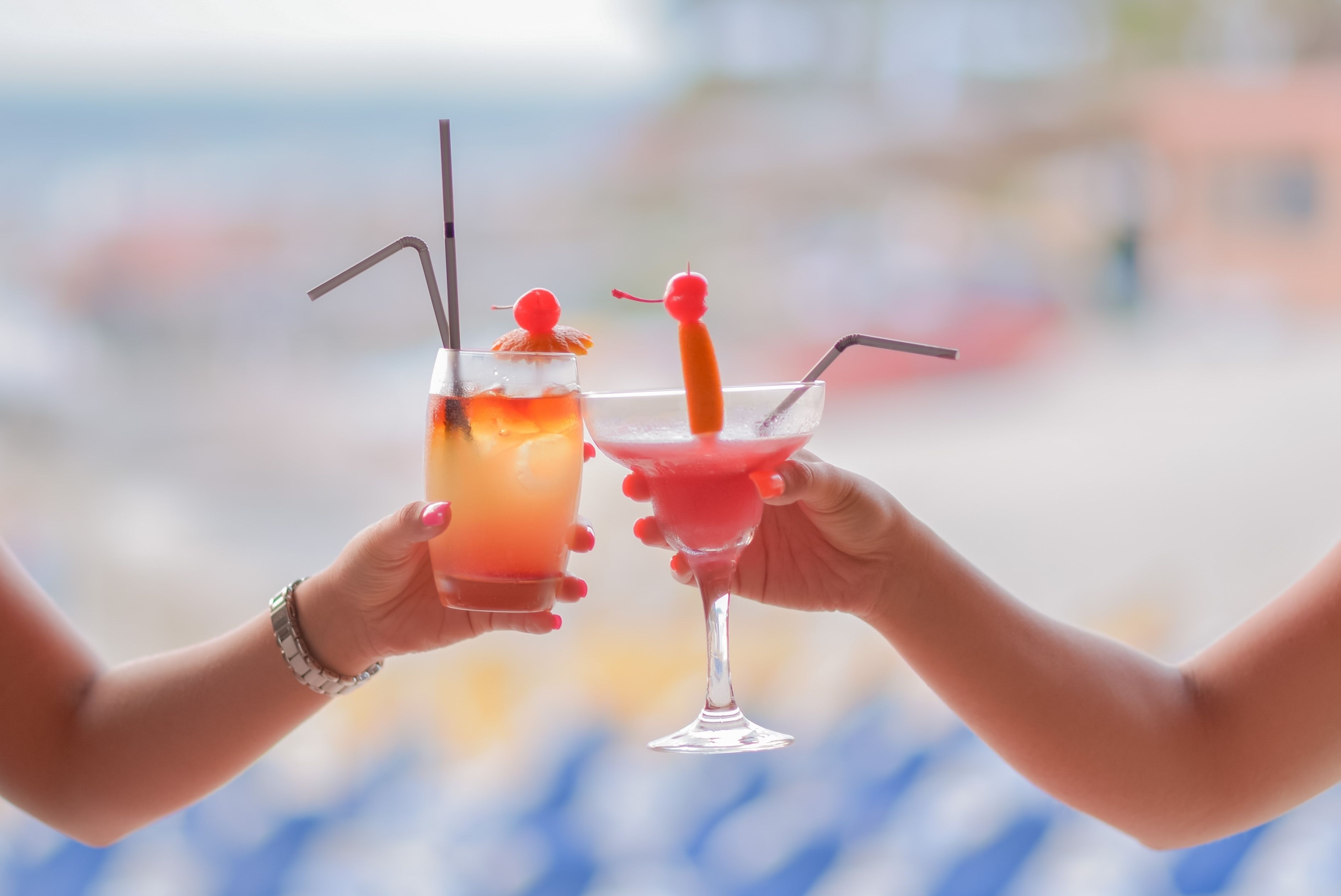 Two hands holding cocktail glasses with straws and cherries.
