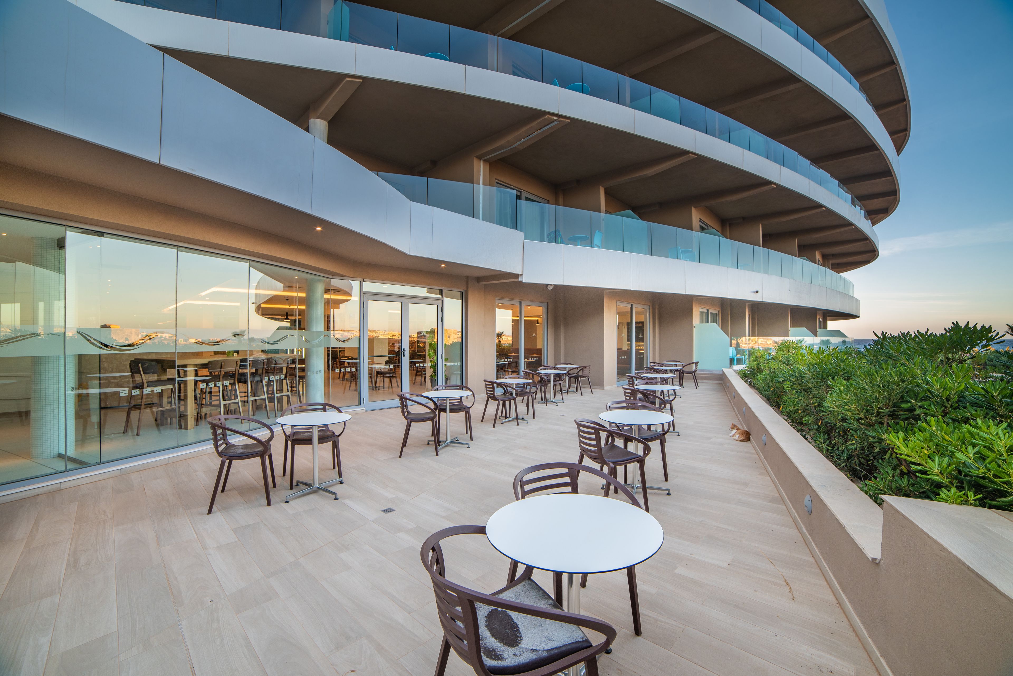 A modern outdoor dining area with multiple tables and chairs, a glass facade, and a curved architectural design.