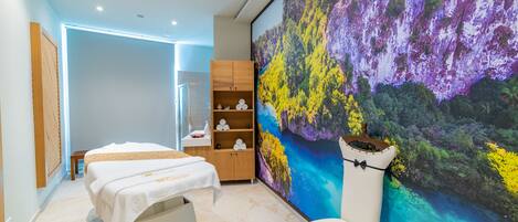 A spa room with a massage table, a wooden cabinet with towels, and a large wall mural of a forest scene.