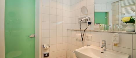 A bathroom with a white sink, a mirror, a hairdryer on a wall-mounted shelf, a toilet, and a green-tinted shower door.