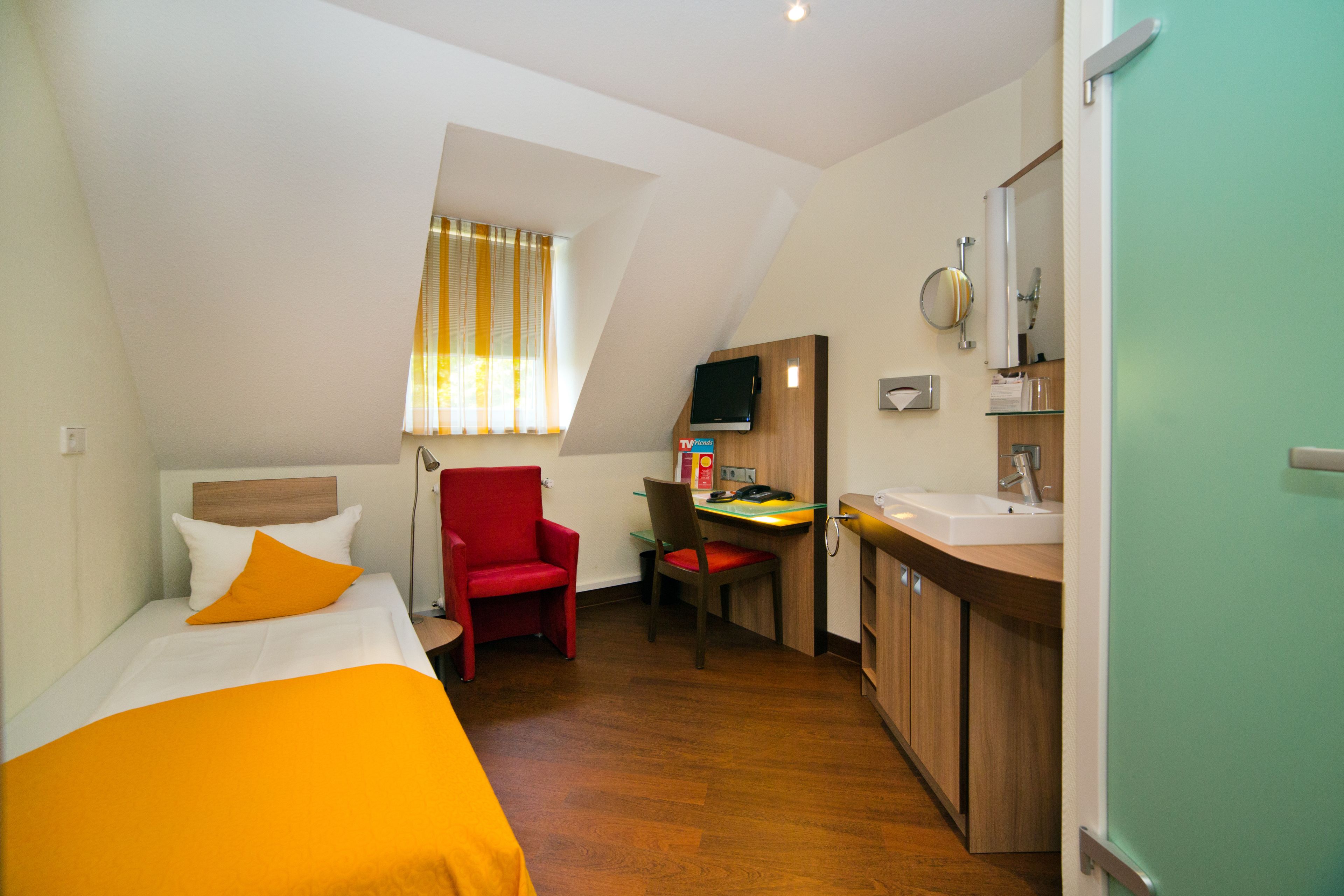 Ein kleines Hotelzimmer mit Bett, Schreibtisch, einem roten Sessel und einer Küchenzeile.