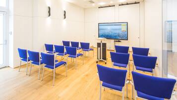 A conference room with blue chairs arranged in rows, a large screen on the wall, and a wooden floor.