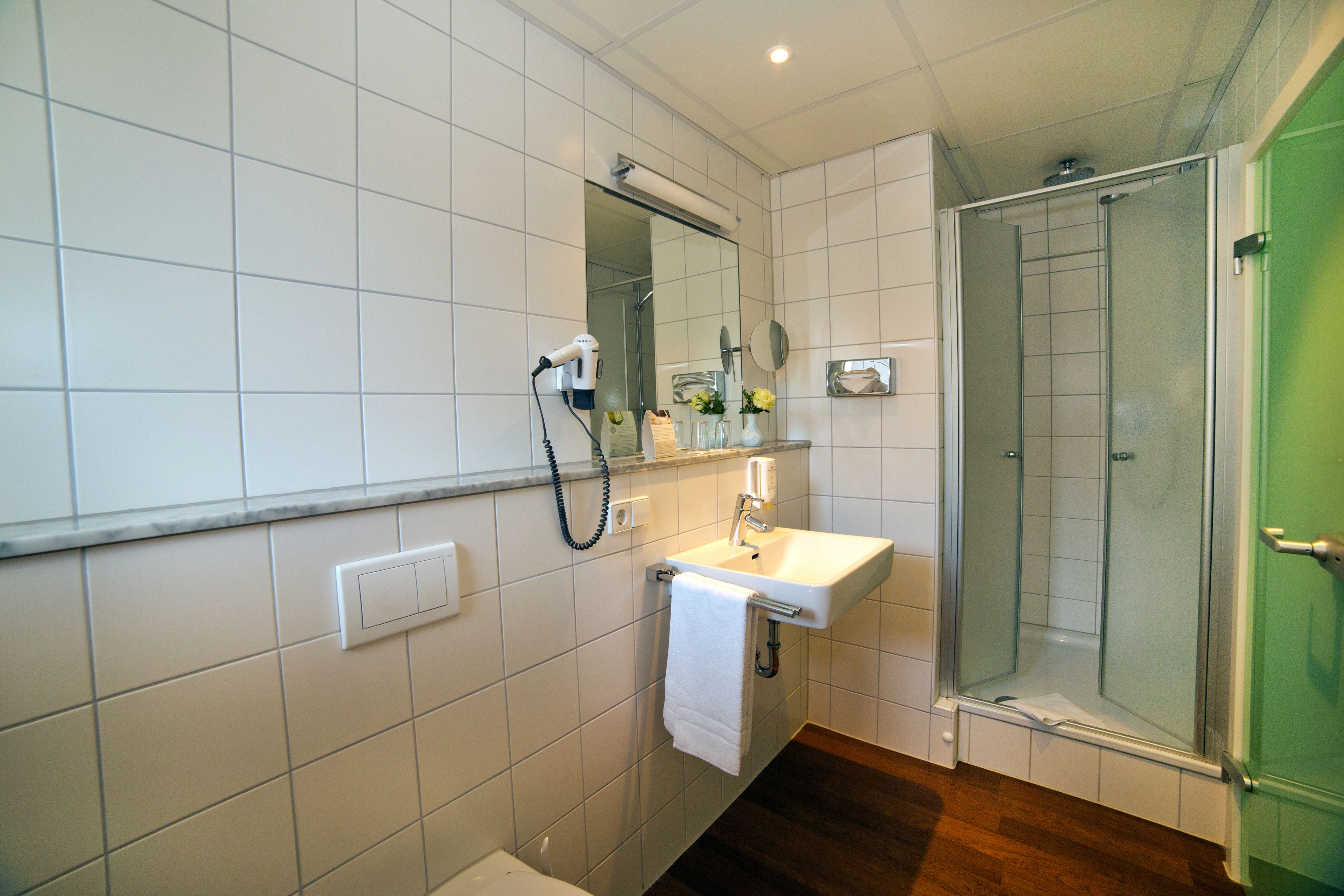 A bathroom with white tiles, a sink, a mirror, a towel rack, a shower with a glass door, and a green door.